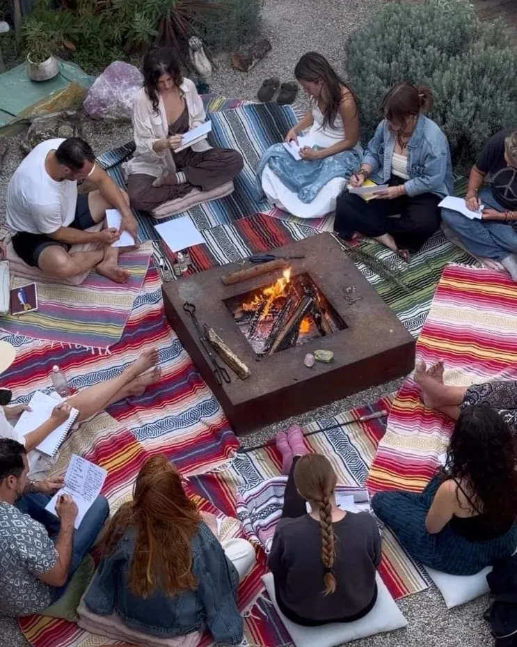 A circle of people sitting on blankets around a fire, writing during an outdoor gathering